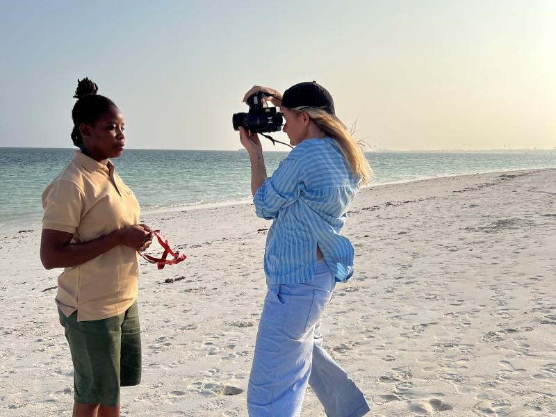Kleis, en hvis kvinde, fotograferer en sort kvinde, de står begge på en strand med hav og himmel bagved.
